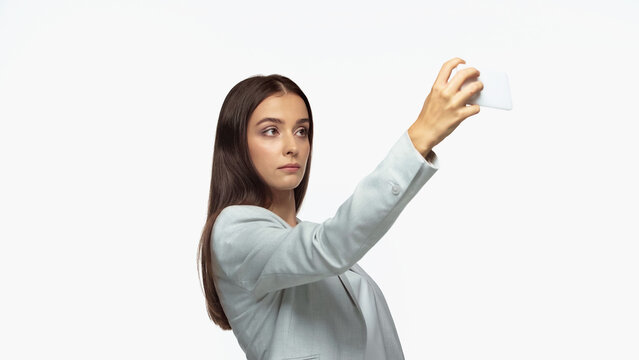 successful businesswoman in grey blazer taking selfie on smartphone isolated on white.