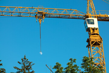 Construction crane on the background of the blue sky and the Moon. Green tree tops