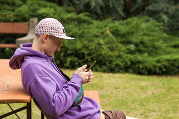 Young caucasian man wears a violet sweatshirt and cap sitting on park bench and using smartphone....