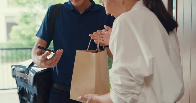 Close-up Young Asian delivery man in blue navy uniform carry case box knock door home online shopping to woman in front of the door and lady sign accept paper bag on smartphone at house concept.