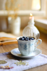 Fototapeta na wymiar Vintage porcelain cup filled with fresh blueberries, pressed flowers, open book, reading glasses and lit candle on the table. Hygge at home. Selective focus.
