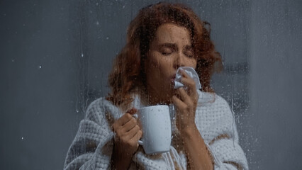 sick woman holding cup with hot beverage and coughing behind window glass with raindrops.