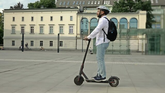 Bearded man with a backpack and in a protective helmet rides an electric scooter through the city square. The concept of convenient and environmentally friendly individual urban transport.