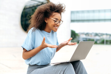 Busy positive young african american woman with glasses, mixed race female sitting outdoors, emotionally talking on video conference using laptop, gesturing with hands, looking at screen, smiling