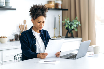 Positive clever young adult african american curly haired woman, freelancer, auditor, working from home, sitting in the kitchen, using a laptop, focused studying financial documents, graphs, reports