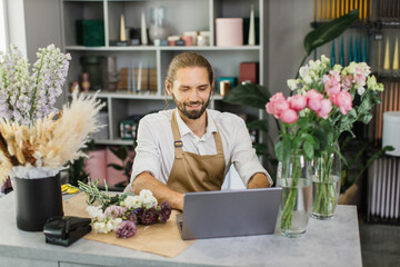 Portrait of attractive man florist working on computer at workplace. Concentrated bearded male sitting at counter in floral store and typing on laptop.