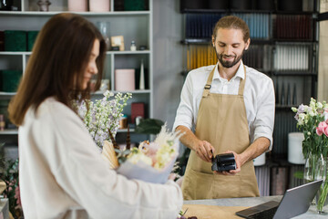 Attractive young man receives payment for a bouquet of flowers with credit card in flower shop. Female customer paying with card for her purchase flowers. Shopping in garden center, business.