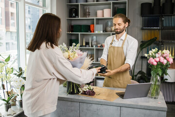 Caucasian female using electronic credit card on her smartphone paying for purchase using payment terminal in flower shop. Client buying bouquet making payment in store - Powered by Adobe
