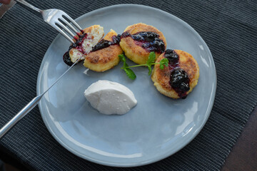 Delicious fried cheesecakes with a crispy crust, poured with cherry jam, served on a gray ceramic plate with a ball of sour cream and decorated with a sprig of fragrant mint