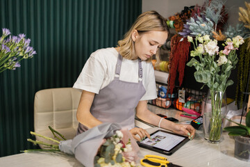 Portrait of caucasian woman entrepreneur sitting in own flower shop, typing on tablet. Young male employee in floral store working on computer. Florist concept.