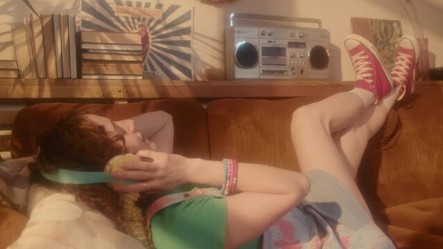 Full side view of red-haired Caucasian girl in over-ear headphones listening to music via tape player, lying on couch in living room at daytime, having fun