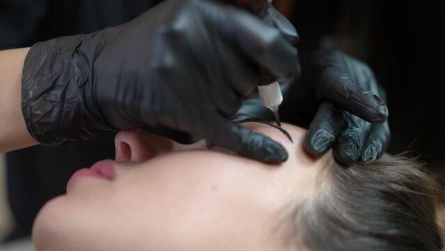 Micro blading tattooing of female eyebrow with microblading pen in beauty salon. Close-up face of Caucasian woman lying with professional tattoo master doing permanent makeup