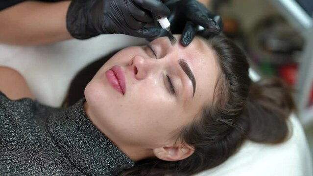 High angle view portrait of Caucasian woman doing permanent makeup with unrecognizable beautician tattooing eyebrow with microblading pen. Beautiful slim client in beauty salon indoors
