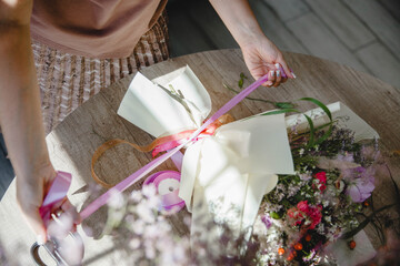 a woman florist ties a pink ribbon bouquet wrapped in paper. The bouquet lies on a round wooden...