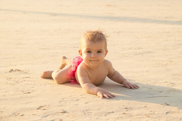 Fototapeta na wymiar Little baby girl is lying on a sandy beach near to sea in sunset sunlight.