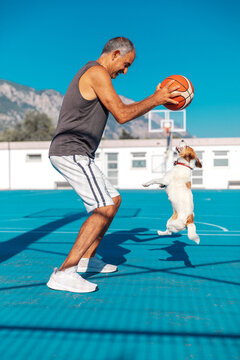 Portrait Of Senior Adult Turkish Cypriot Man Playing Basketball With A Small Cute Jumping Dog Jack Russel Terrier On Playground Outdoor At Sunny Summer Day.