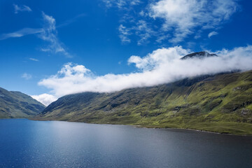 Fototapeta premium Green mountains under blue cloudy sky on a sunny warm day and lake with blue water. Nature scene in Connemara, Ireland. Irish landscape. Popular travel and sightseeing area.