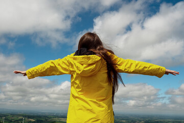 Teenager girl in yellow jacket with hands up in the air. White cloudy sky background. Positive body language. Copy space.