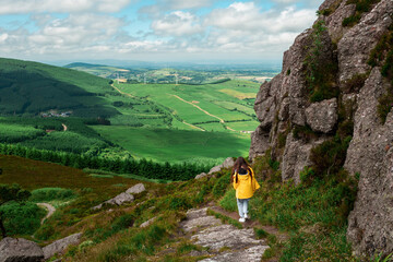 Teenager girl walking on a foot path in a mountain. Stunning green nature background with fields and wind power generators and blue cloudy sky. Hiking and outdoor activity concept. Tipperary, Ireland
