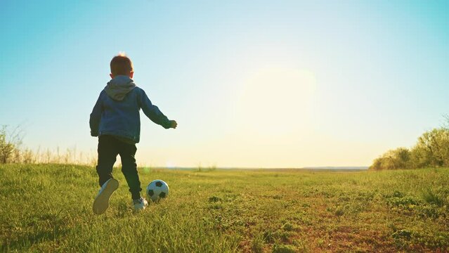 cute little boy is playing football in field in nature in sunny summer day, slow motion, funny preschooler child is running and kicking soccer ball, happy childhood moments