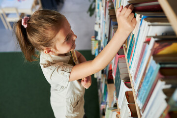 School girl looking at bookshelf in school library. Smart girl selecting literature for reading....