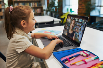 Student litttle girl having video class remotely with her teacher on laptop sitting at desk in...