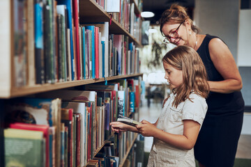 Teacher helping to choose book her schoolgirl in school library. Smart girl selecting literature...