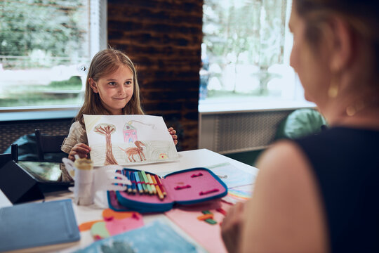 Girl presenting her artwork teacher. Woman assisting schoolgirl during classes at primary school. Child drawing picture sitting at desk in classroom. Learning at primary school. Back to school