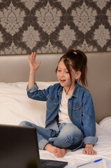 Happy cute little girl study online sitting on her bed with a laptop and notebooks. Schoolgirl has a video call and rises her hand in her bedroom. Distance education concept