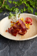 Maple Syrup glazed bacon over caramelized corn with sprout garnish and cherry tomato on a compostable bamboo plate