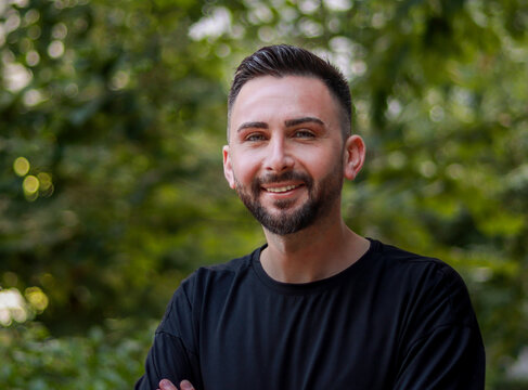 Portrait of young handsome man with beard. Young man in black shirt smiling looking at camera. Outdoor nature background