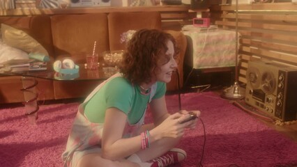 High angle of ginger Caucasian girl in outfit in 80s fashion on floor in living room at daytime, playing 8-bit computer game, using gamepad and console