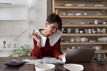 Female artisan running small business analyzing bills and receipts while standing at table in ceramic art studio, doing pottery cost analysis, calculating fixed and variable costs of small business