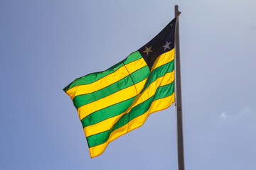 Uma bandeira do estado de Goiás hasteada e balançando ao vento com céu ao fundo.