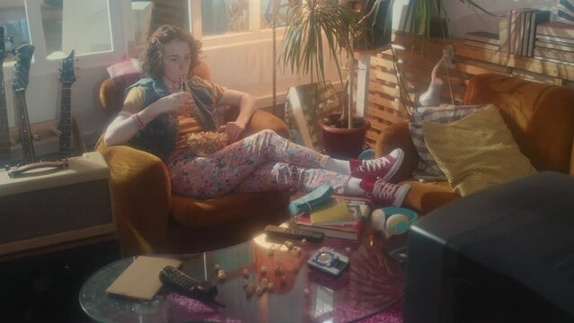 Long shot of young Caucasian girl sitting in armchair in room with interior design in eighties style, watching movie on retro TV, drinking soda and eating popcorn