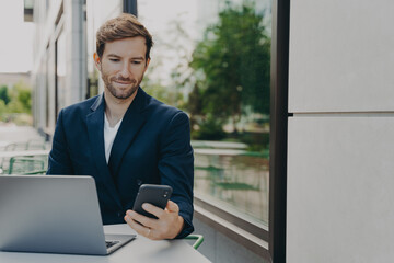 Fototapeta premium Focused young businessman working on laptop in outdoor cafe and browsing phone notifications