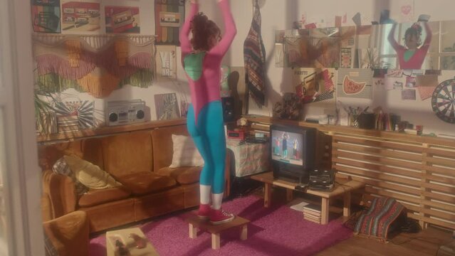 Rear full shot of red-haired Caucasian girl in eighties aerobic outfit exercising with fitness tape running on retro TV in living room in afternoon