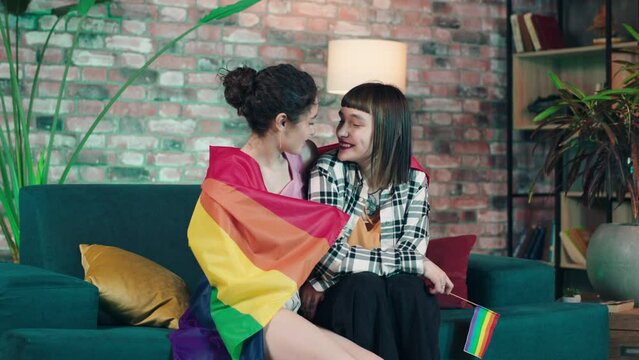 Happy lesbian couple together sitting on the sofa in living room and enjoy the time they holding a LGBT rainbow flag and smiling cute to each other