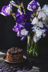 Tasty chocolate cake with blackberries decoration and beautiful bouquet of iris flowers in vase on table. Blooming lilac and white flowers. Present for birthday. Summertime gift. 