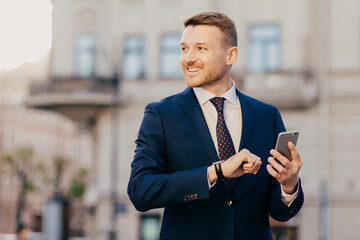 Horizontal shot of successful unshaven male CEO looks for interesting multimedia files on smart phone, downloads or updates new programs, stands against blurred city background, looks happily aside