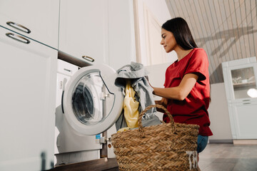 Young woman putting clothes at washing machine while doing laundry