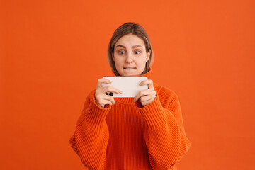 Young beautiful tense girl playing mobile game on her phone standing over isolated orange background