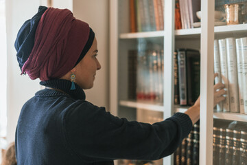 Religious jewish young woman with head covered stands near bookcase with religious books (9) - obrazy, fototapety, plakaty