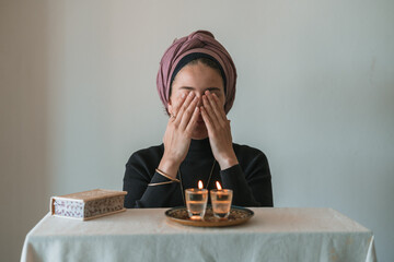 Jewish woman prays over lit Shabbat candles, covering her face with her hands. Nearby lies a...