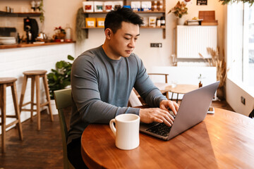 Serious adult asian man using laptop while sitting in cafe indoors