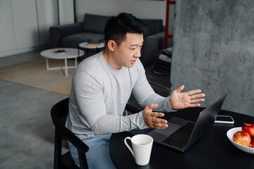 Asian man gesturing while working with laptop in kitchen