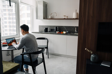 Asian man wearing glasses working with laptop in kitchen