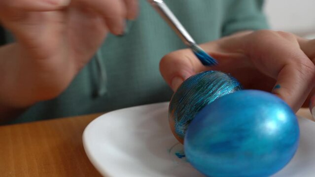 A young girl with a brush paints a white chicken egg blue on a white plate in honor of the Easter holiday. Close up