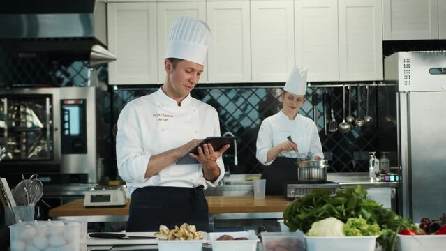 Restaurant kitchen: portrait of male and female cooks. Male Chef checks the availability of products in the warehouse using a tablet