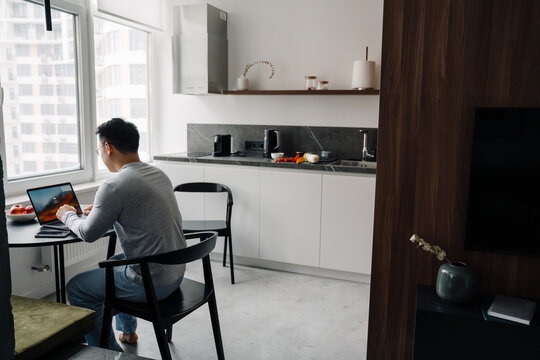 Asian Man Wearing Glasses Working With Laptop In Kitchen